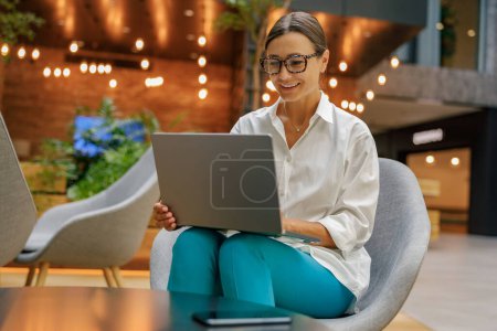 Foto de Sonriendo mujer de negocios elegante que trabaja en el ordenador portátil sentado en el fondo moderno de la oficina - Imagen libre de derechos
