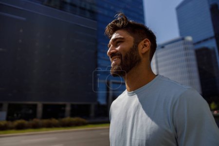 Téléchargez les photos : Vue arrière de l'homme souriant barbu en t-shirt debout sur fond de gratte-ciel. Photo de haute qualité - en image libre de droit