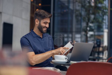 Téléchargez les photos : Souriant beau gestionnaire masculin regarde le bloc-notes tout en travaillant sur ordinateur portable dans le coworking confortable - en image libre de droit