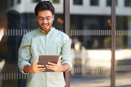 Téléchargez les photos : Joyeux freelance dans les lunettes de travail sur tablette numérique debout près du bâtiment de coworking - en image libre de droit