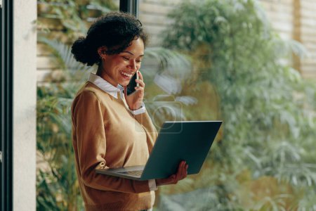 Téléchargez les photos : Femme pigiste parle téléphone tout en travaillant sur ordinateur portable de la maison debout près de la fenêtre - en image libre de droit