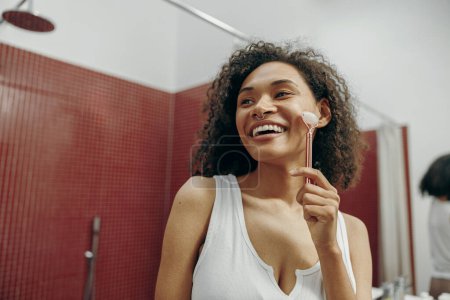 Téléchargez les photos : Femme heureuse à la salle de bain utiliser rouleau facial pour les soins de la peau et regarde ailleurs. Concept de soins de la peau - en image libre de droit