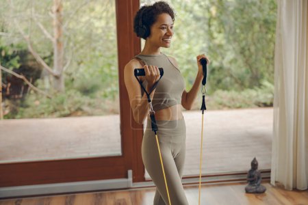 Photo for Smiling woman in sportswear exercising with resistance band standing on yoga mat at home - Royalty Free Image