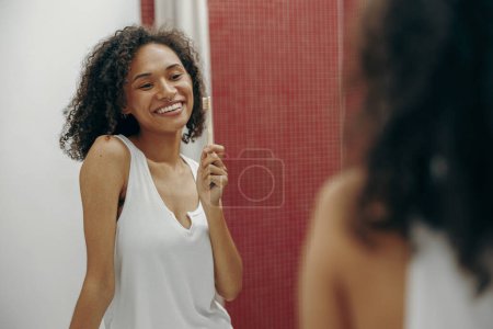 Téléchargez les photos : Jeune femme brossant les dents avec une brosse à dents le matin et regardant dans le miroir dans la salle de bain - en image libre de droit