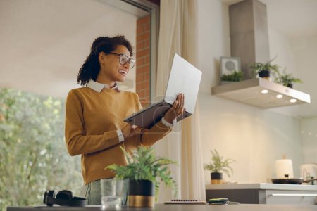Téléchargez les photos : Attrayant femme d'affaires travaillant à distance sur ordinateur portable de la maison tout en se tenant près de la fenêtre - en image libre de droit
