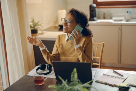Téléchargez les photos : Femme souriante parle smartphone tout en se tenant à la maison tout en travaillant sur un ordinateur portable et regarde ailleurs - en image libre de droit