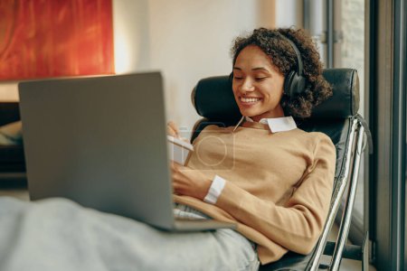 Téléchargez les photos : Jeune étudiante souriante étudiant sur un ordinateur portable et prenant des notes tout en étant allongé sur une chaise à la maison - en image libre de droit