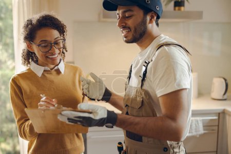Téléchargez les photos : Sourire jeune femme signant document tout en communiquant avec bricoleur à la cuisine - en image libre de droit