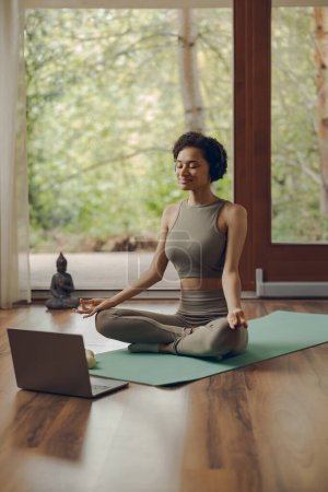Foto de Mujer con auriculares practicando yoga y escuchando meditación en casa sentada en pose de loto sobre alfombra - Imagen libre de derechos