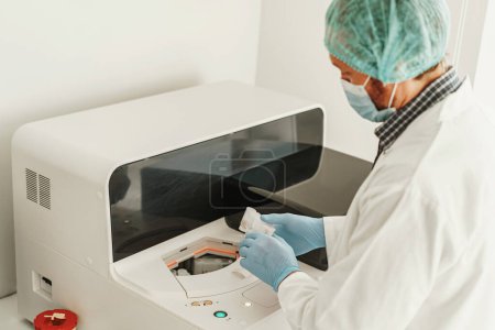 Foto de Científico masculino en uniforme y máscara poniendo un pequeño tubo de ensayo de plástico en una microcentrifugadora - Imagen libre de derechos
