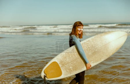 Foto de Surfista femenina en traje de neopreno con su tabla de surf entrando fuera del mar después de surfear en las olas - Imagen libre de derechos