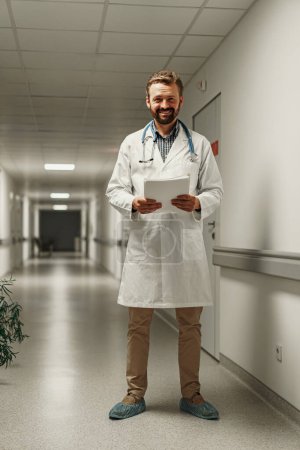 Foto de Médico varón vestido de blanco con estetoscopio de pie en la sala de la clínica y la celebración de documentos en las manos - Imagen libre de derechos