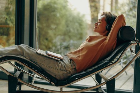 Foto de Smiling freelancer femenina en auriculares está descansando mientras está acostado en la silla durante el recreo en casa - Imagen libre de derechos