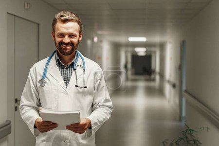 Photo for Doctor standing at hospital hall and holding clipboard with patient records looks at camera - Royalty Free Image