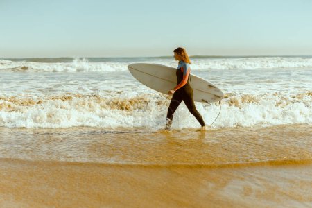 Foto de Surfista femenina en traje de neopreno con su tabla de surf entrando en el mar. Surf y océano. Foto de alta calidad - Imagen libre de derechos