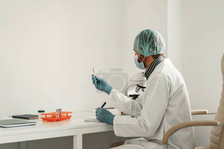 Foto de Hombre científico en máscara protectora toma notas en informe sentado en el laboratorio con microscopio - Imagen libre de derechos