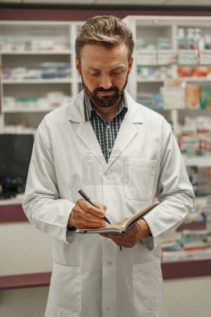 Photo for Male pharmacist in uniform is making notes in notepad while work in pharmacy. High quality photo - Royalty Free Image