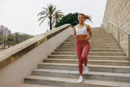 Photo for Smiling active female athlete in sportswear running on steps outdoors in early morning - Royalty Free Image
