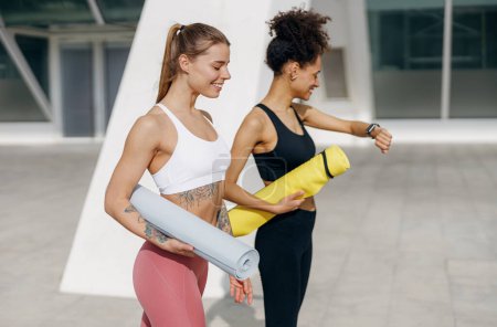 Foto de Dos amigas sonrientes caminando sobre el fondo matutino de la ciudad con colchonetas de yoga después de hacer gimnasio matutino - Imagen libre de derechos