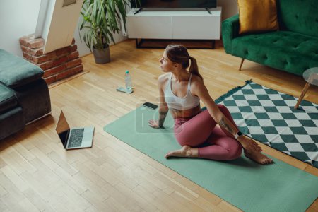 Téléchargez les photos : Femme sur tapis de yoga en vêtements de sport est assis en face de l'ordinateur portable sur le sol et les jambes tendues - en image libre de droit