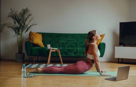 Foto de Una mujer está practicando yoga en una alfombra colocada en pisos de madera frente a un portátil - Imagen libre de derechos