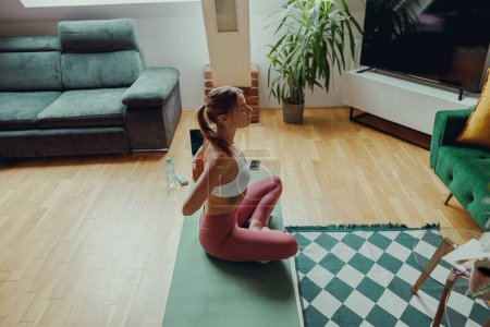 Foto de Una mujer está sentada en una esterilla de yoga en una sala de estar con pisos de madera, rodeada de plantas - Imagen libre de derechos