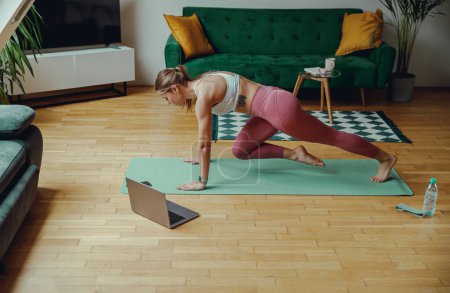 Téléchargez les photos : Jeune femme en tenue de sport tient une position de planche sur un tapis de yoga dans un salon - en image libre de droit