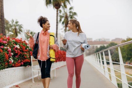 Foto de Dos mujeres jóvenes en forma caminando por la ciudad con sus esterillas de yoga después de hacer ejercicio matutino - Imagen libre de derechos