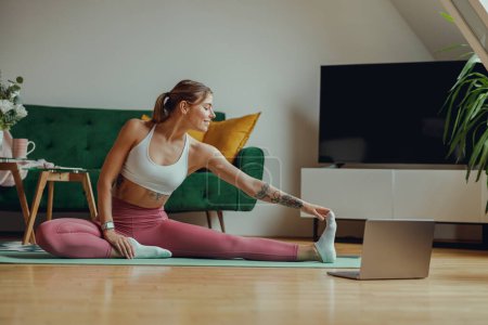 Photo for A woman is sitting on a yoga mat on a hardwood floor and stretching her legs. High quality photo - Royalty Free Image