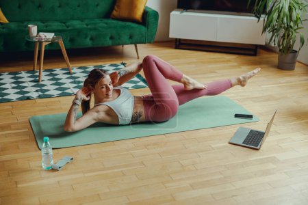 Photo for A woman in an undershirt is doing exercises on a yoga mat on the hardwood flooring of a living room - Royalty Free Image