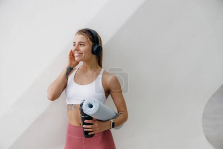 Téléchargez les photos : Athlète féminine souriante écoutant de la musique dans un casque tout en se reposant après avoir fait du sport à l'extérieur - en image libre de droit