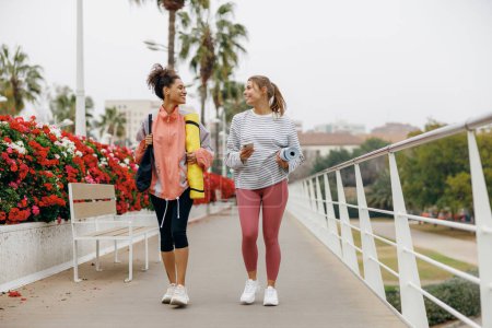 Photo for Two young fit women walking in the city with their yoga mats after doing morning workout - Royalty Free Image