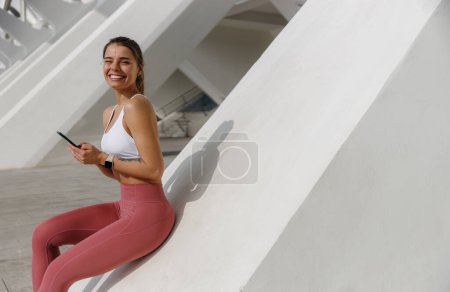 Photo for Smiling woman in sportswear sitting outdoors after training and use mobile phone and looks camera - Royalty Free Image