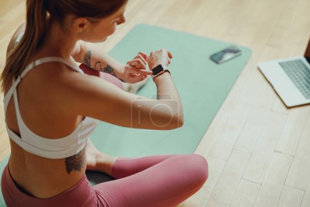 Téléchargez les photos : Une femme s'assoit sur un tapis de yoga et regarde sa montre intelligente avec un ordinateur portable sur le sol - en image libre de droit