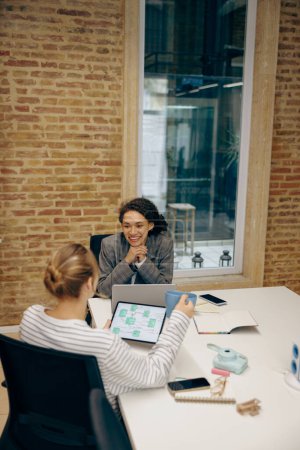 Foto de Dos mujeres freelancers sonrientes hablando durante el trabajo en el portátil mientras están sentadas en un acogedor coworking - Imagen libre de derechos