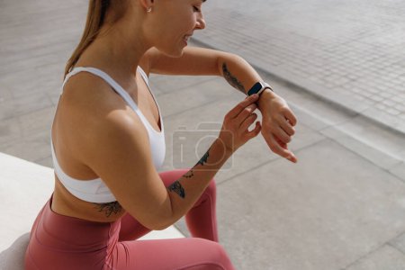 Téléchargez les photos : Femme souriante en tenue de sport regardant smartwatch après avoir couru. Sports de plein air le matin - en image libre de droit