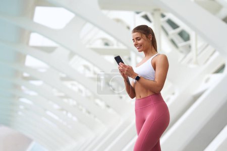 Photo for A woman in a shoulderbaring white tank top and pink leggings is elegantly looking at her phone - Royalty Free Image