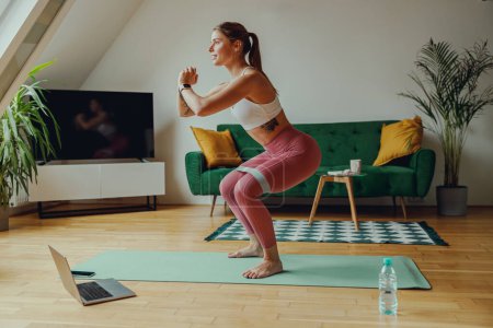 Foto de Una mujer está en cuclillas en una estera de yoga en una sala de estar con pisos de madera - Imagen libre de derechos