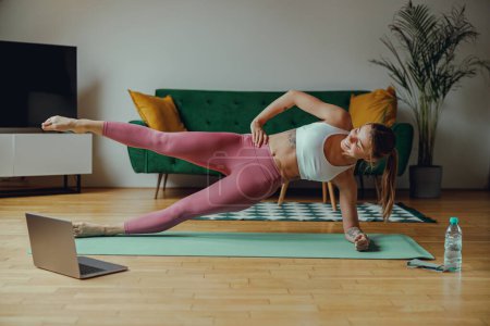 Photo for Young woman in sportswear is holding a plank position on a yoga mat in a living room - Royalty Free Image