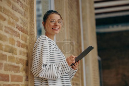 Téléchargez les photos : Jeune femme souriante pigiste tenant tablette numérique et regarde la caméra debout dans le coworking - en image libre de droit