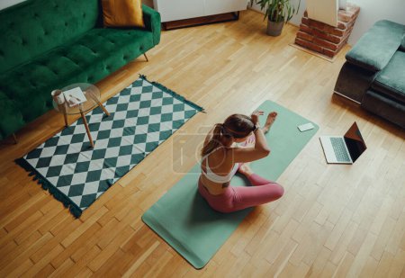 Foto de Una mujer está sentada en una esterilla de yoga en el suelo de madera en la sala de estar - Imagen libre de derechos