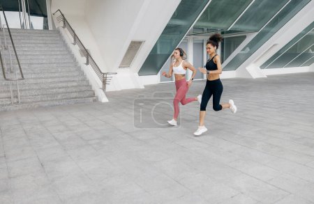 Téléchargez les photos : Deux jeunes femmes en tenue de sport courent sur fond de bâtiments modernes. Concept de mode de vie actif - en image libre de droit