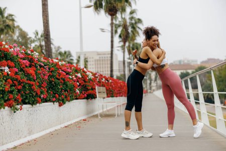 Téléchargez les photos : Des femmes souriantes et gaies s'embrassent en vêtements de sport marchant après l'exercice en ville - en image libre de droit
