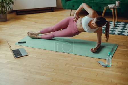 Photo for Young woman in sportswear is holding a plank position on a yoga mat in a living room - Royalty Free Image
