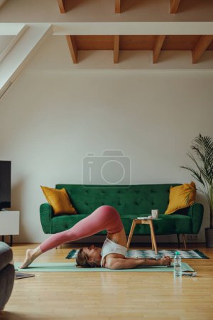 Photo for A woman is performing a yoga pose on a wood floor in a living room, with her leg extended - Royalty Free Image