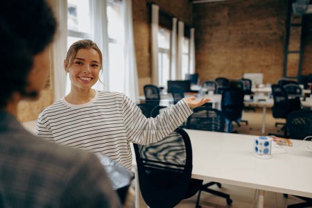 Pretty smiling female entrepreneur doing welcome gesture to colleague while standing in office