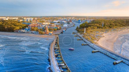 Photo for Drone photo captures Kolobrzeg's maritime charm, featuring the iconic lighthouse, cerulean sea, turbulent waves, a distant pier, and autumnal hues on the trees. - Royalty Free Image