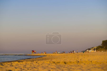 Photo for A beach in Mrzezyno, West Pomeranian Voivodeship, Poland, captured during the golden hour, beautifully illuminated by warm sunlight. People strolling and sunbathing, clear sky, vegetation on dunes, stairs leading to the beach. - Royalty Free Image