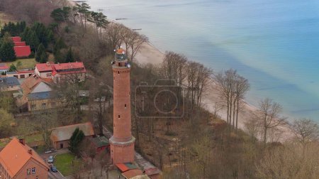 A drone shot displays Gaski beach, West Pomeranian Voivodeship, Poland, with a red brick lighthouse, Baltic Sea, sandy beach, leafless dune trees, holiday cottages, hotels, and homes. Possibly calm sea. Captured in February winter.