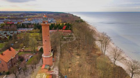 Eine Drohnenaufnahme zeigt den Strand Gaski in der Woiwodschaft Westpommern, Polen, mit einem Leuchtturm aus rotem Ziegelstein, Ostsee, Sandstrand, grünen Dünenbäumen, Ferienhäusern, Hotels und Häusern. Möglicherweise ruhige See. Im Februar-Winter gefangen genommen.